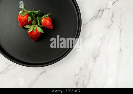 Draufsicht auf schwarzes Schild auf weißem Marmorhintergrund. Drei Erdbeeren lagen flach. Speicherplatz kopieren. Stockfoto