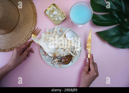 Kreative Fotografie. Das Konzept der Sommerferienerinnerungen. In den Händen der Frauen sind goldene Essutensilien, auf dem Tisch ist ein Strohhut, ein Laken Monstera und ein Teller mit Muscheln. Stockfoto