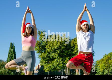 Bild eines Paares, das mit dem Weihnachtsmann und Sportswear in the Park Yoga zu Weihnachten macht. Der Mann genießt es und die Frau hasst den Sport Stockfoto