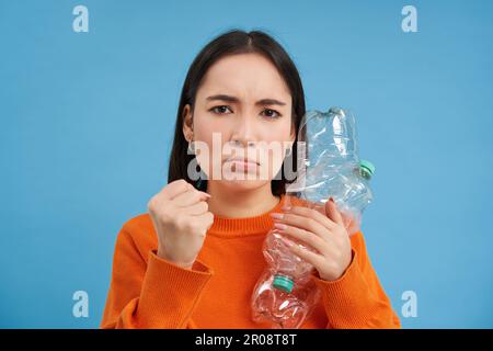 Wütende asiatische Frau mit zwei Plastikflaschen mit Stirnrunzeln, Mädchen mit umweltfreundlichem Lebensstil, Drang, Abfall zu recyceln, blauer Hintergrund Stockfoto