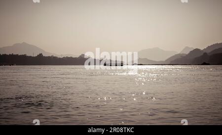 Sepiabarbene Silhouette des Slow Bootes auf dem Mekong, der an Laos und Thailand grenzt, mit Umrissen der umliegenden Berge. Stockfoto