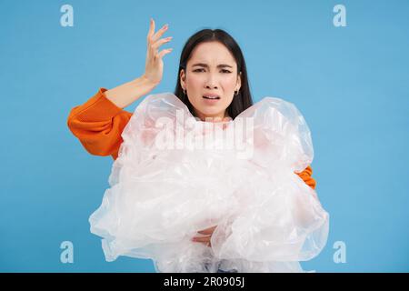 Genervte asiatische Freiwillige, die Plastikabfall in der Hand hält, mit enttäuschtem Gesicht schreit, die Verschmutzung hasst, steht über blauem Hintergrund Stockfoto