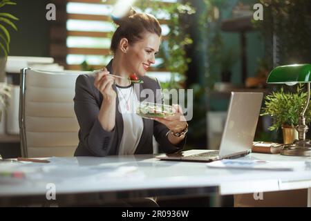 Nachhaltiger Arbeitsplatz. Glückliche, moderne 40 Jahre alte Buchhalterin in einem grauen Business-Anzug in einem modernen grünen Büro mit Laptop-Esssalat. Stockfoto