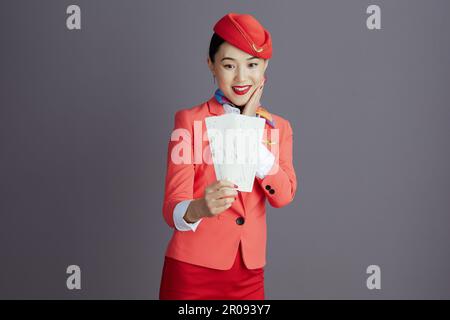 Lächelnde, stilvolle asiatische Stewardess in rotem Rock, Jacke und Hut Uniform mit Flugtickets vor grauem Hintergrund. Stockfoto