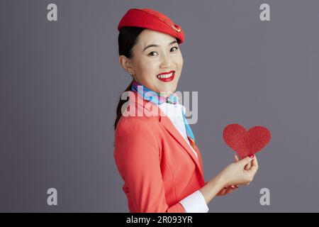 Lächelnde, stilvolle asiatische Stewardess in rotem Rock, Jacke und Hut Uniform mit rotem Papierherz vor grauem Hintergrund. Stockfoto