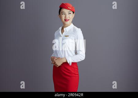 Eine nachdenkliche, elegante Stewardess, asiatische Frau mit rotem Rock und Hut Uniform, die auf den Kopierbereich vor grauem Hintergrund blickt. Stockfoto