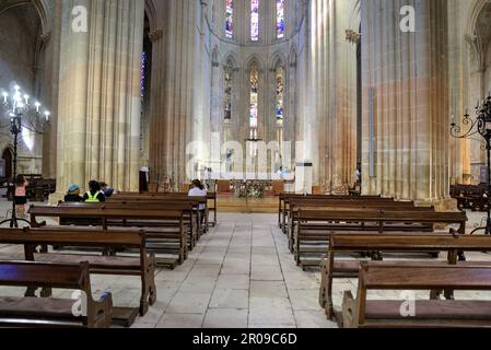 Batalha, Portugal - 15. August 2022: Das Innere des gotischen Klosters zeigt einen Altar mit Kreuz, Buntglasfenster, Bänke und Touristen Stockfoto