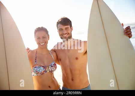 Wir sind in der Brandung. Porträtaufnahme eines glücklichen jungen Paares, das mit seinen Surfbrettern am Strand lächelt. Stockfoto