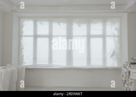 Fenster mit Plastikfolie und Baumaterialien im renovierten Zimmer Stockfoto