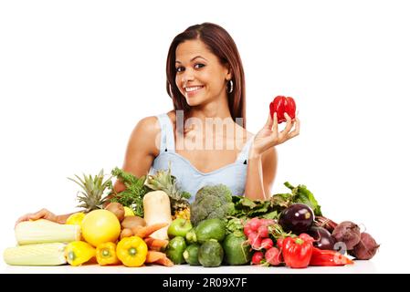 Sie liebt gesundes Essen. Attraktive junge Frau lächelt, während sie einen roten Pfeffer neben einem Haufen frisches Gemüse auf weißem Hintergrund hält. Stockfoto