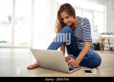 Was soll ich mir heute ansehen? Ganzkörperaufnahme einer attraktiven jungen Frau, die tagsüber im Wohnzimmer mit einem Laptop sitzt. Stockfoto