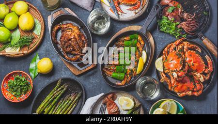 Tisch mit Meeresfrüchtegerichten - gekochte Krabben, Tigergarnelen, gegrillte Tintenfische und Tintenfische auf gusseisernen gegrillten Pfannen und Tellern, Weißwein. Getöntes Foto Stockfoto