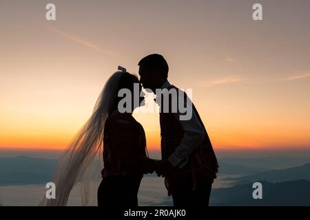 Silhouette eines verliebten Ehepaars, das sich küsst und bei Sonnenaufgang Hand an Hand hält, mit morgendlichem Himmelshintergrund Stockfoto