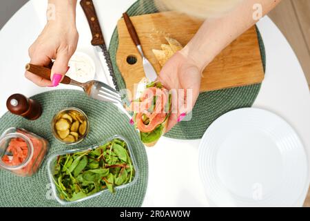 Draufsicht der Hände einer Frau mittleren Alters legt geräucherte Forelle auf ein Sandwich, während sie Frühstück macht Stockfoto