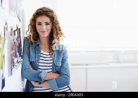 Eine Karriere im Grafikdesign zu verfolgen. Eine junge Grafikdesignerin lächelt die Kamera an, während sie neben einer Anordnung von Fotos an einer Wand steht. Stockfoto