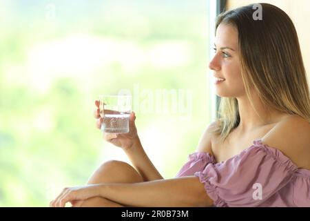 Glücklicher Teenager mit einem Glas Wasser blickt durch ein Fenster zu Hause oder im Hotel Stockfoto