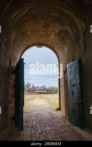 Zwei schwere dunkelgrüne Türen stehen am Ende eines gewölbten Ziegeltunnels offen und rahmen ein entferntes Ziegelgebäude im Fort Washington Park, Maryland, ein. Stockfoto