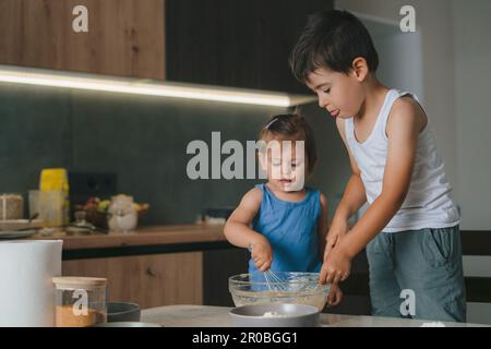 Mädchen und Bruder mischen Teigzutaten in einer Schüssel mit Schneebesen. Zukünftige Koch- und Kinderspiele in der Küche. Lebensmittelzubereitung. Glückliche Familie. Stockfoto