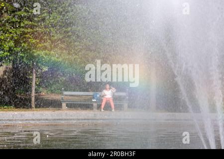 Eine Dame sitzt auf einer Bank im Parc de Bruxelles, durch einen Brunnen mit Regenbogeneffekt gesehen. Stockfoto