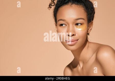 Eine afroamerikanische Frau mit goldenen Hydrogel-Pflastern unter den Augen, um Schwellungen bei kosmetischen Eingriffen zu lindern, isoliert auf beigem Hintergrund. Haut Stockfoto