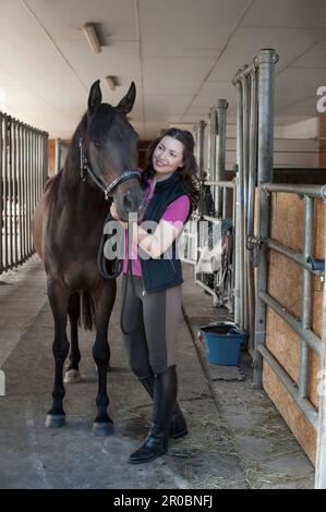 Reife Frau streichelt ihr braunes Pferd im Stall und lächelt, Bayern, Deutschland Stockfoto