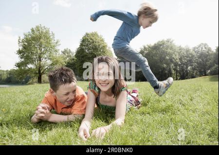 Ein Junge, der über seine Freunde springt, liegt auf Gras in einem Park, München, Bayern, Deutschland Stockfoto
