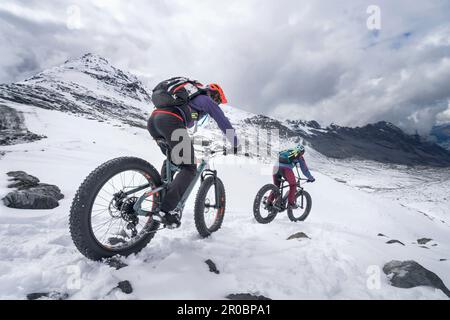 Paar reiten Elektrische fatbikes auf schneebedeckte Berge, Passo di Stelvio, Lombardei, Italien Stockfoto