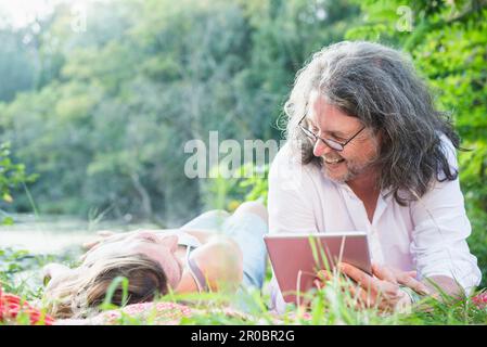 Ein Paar, das Seite an Seite mit einem digitalen Tablet am See liegt, Deutschland Stockfoto