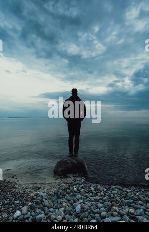 Silhouette eines Mannes, der in der Dämmerung allein auf einem Felsen im Wasser steht. Stockfoto