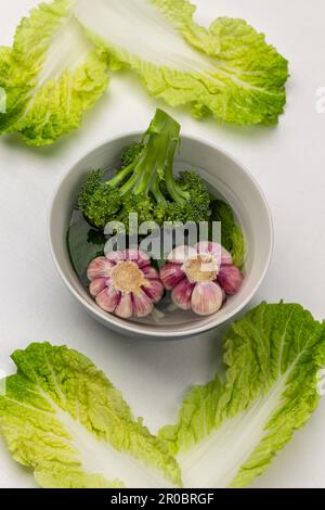 Zwei Köpfe Knoblauch und Brokkoli in einer Schüssel Wasser. Gelbgrüne Salatblätter. Flach verlegt. Weißer Hintergrund. Stockfoto