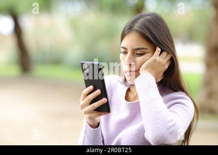 Eine gelangweilte Frau überprüft ihr Smartphone, wenn sie in einem Park sitzt Stockfoto