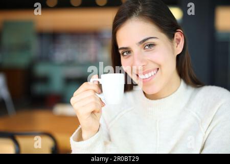 Glückliche Frau, die lächelt und eine Kaffeetasse in einer Bar hält und in die Kamera schaut Stockfoto