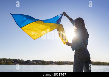 Weibliche Silhouette hält eine ukrainische Flagge, die im Wind flattert, wunderschön von der Sonne beleuchtet Stockfoto