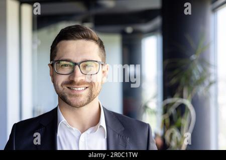 Nahaufnahme Foto eines jungen erfolgreichen Unternehmertums, Geschäftsmann mit Brille am Arbeitsplatz, der lächelt und in die Kamera schaut. Stockfoto