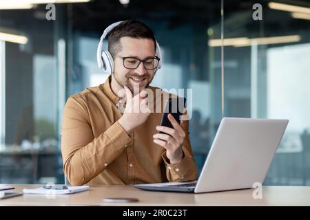 Junger Programmierer, der im Büro mit einem Laptop arbeitet, Mann im Kopfhörer, der lächelt und Online-Musik hört, Audio-Podcast und Hörbuch, Geschäftsmann im Hemd am Arbeitsplatz, der ein Telefon in der Hand hält. Stockfoto