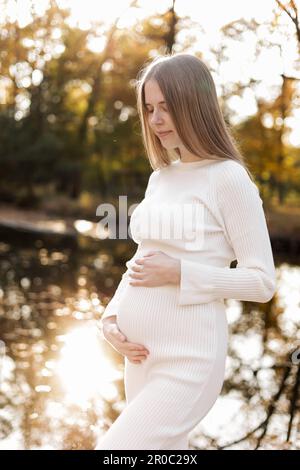 Muttertag. Wunderschöne junge schwangere Frau in weißem, eng gestricktem Kleid mit kleinem Bauch, der sich draußen am Teich mit reflektierender Natur und Sonne i entspannt Stockfoto