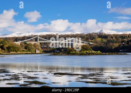 Malerischer Blick auf die Menai-Hängebrücke, die die Menai-Straße überquert, mit Schnee auf den Bergen im Winter. Menai Bridge (Porthaethwy), Anglesey, Wales, Vereinigtes Königreich Stockfoto