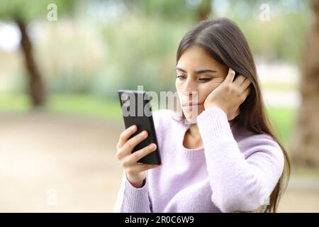 Eine gelangweilte Frau überprüft ihr Smartphone, wenn sie in einem Park sitzt Stockfoto