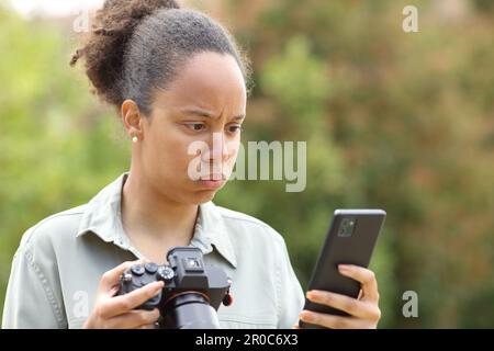 Frustrierter schwarzer Fotograf, der eine spiegellose Kamera hält und das Handy in einem Park überprüft Stockfoto