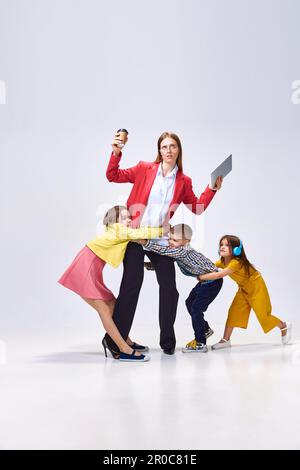 Porträt einer beschäftigten Frau mit Tablet und Kaffee vor grauem Studiohintergrund. Kleine Kinder, die sie umarmen, von der Arbeit stören Stockfoto