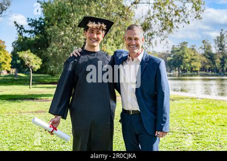 Porträt eines glücklichen kaukasischen jungen Mannes, der an seinem Abschlusstag mit seinem Vater abgeschlossen wurde. Beide blicken auf die Kamera Stockfoto