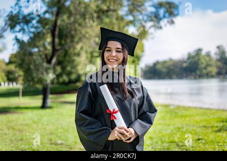 Glücklich kaukasischen graduierten Mädchen mit langen braunen Haaren. Sie trägt ein Junggesellenkleid und ein schwarzes Mortarboard. Sie hat ein Diplom Stockfoto