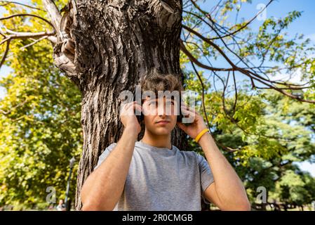 Hübscher junger Mann, der sich auf einen Baum lehnt, während er Musik hört Stockfoto