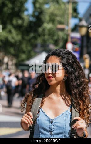 Attraktiver junger türkischer Tourist, der mit einem Rucksack auf der Straße spaziert und wegschaut. Sie trägt ein Jeanskleid und eine Sonnenbrille Stockfoto