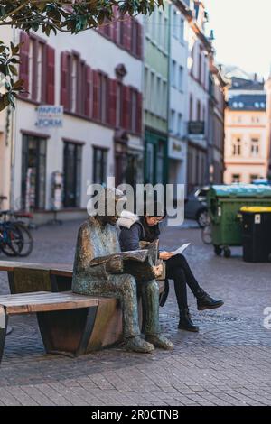 Bronzestatue eines Zeitungslesers. Eine junge Frau sitzt auf einer Bank neben der Statue und liest in Heidelberg Stockfoto