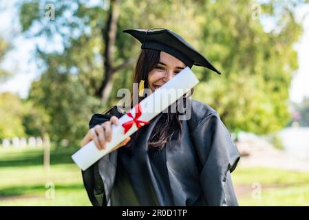 Glückliches, weißes Mädchen mit langem braunem Haar, das ihr Diplom zeigt. Sie trägt ein Junggesellenkleid und ein schwarzes Mörtelbrett. Stockfoto