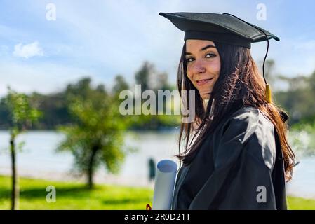 Glückliches, weißes Mädchen mit langem braunem Haar, mit einem Diplom in der Hand und schaut in die Kamera. Stockfoto