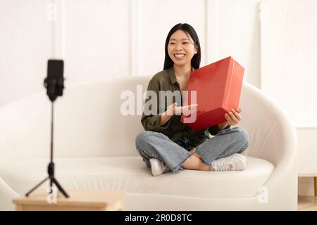 Eine glückliche asiatische Beeinflusserin hält ein rotes Paket und streamt Stockfoto