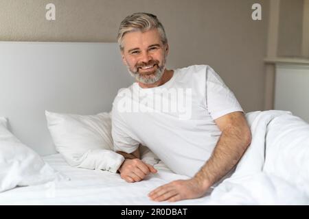 Glücklicher, gutaussehender Mann mittleren Alters, der im Bett sitzt und lächelt Stockfoto