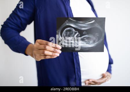 Eine junge afroamerikanische Schwangere mit großem Bauch zeigt ein Ultraschallbild des Babys im Wohnzimmer Stockfoto